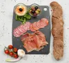 A charcuterie board with various meats, olives, hummus, tomatoes, garlic, and a baguette on a black cutting board. Produktbild-Vorschau