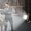 A man is working in a warehouse with stacks of wooden pallets in the background. A portable LED work light is illuminating the scene. Produktbild-Vorschau