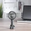 A portable fan is shown on a wooden desk next to a laptop. The background is a blurred office or home environment with shelves and plants. Náhled obrázku produktu