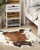 A cow-print rug is placed on a wooden floor next to a white bedside table with wooden drawers and a bed with a white frame and a gray blanket. Podgląd zdjęcia produktu