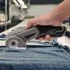 A hand holding a cordless electric rotary cutter over a pile of denim fabric. Náhled obrázku produktu