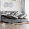 A grey upholstered bed with its storage compartments open, revealing the storage space underneath. The bed is made up with pillows and blankets. Produktbild-Vorschau