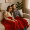 A couple is sitting on a couch, covered by a red blanket, watching TV. Produktbild-Vorschau