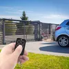 A hand holding a remote control in front of a gate and a car. Voir les images du produit