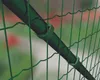 A close-up view of a green fence with a dark green pole attached by green clips. The background is blurred with hints of green foliage and red flowers. Anteprima dell’immagine prodotto