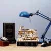 A wooden marble run model and a blue desk lamp are displayed on a wooden surface, with books and a small gear in the foreground. Náhľad obrázku produktu
