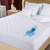 A white quilted mattress protector on a bed, with a glass of blue liquid spilled on it. Produktbild-Vorschau