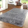 A fluffy gray rug is placed on a wooden floor in a room with a window, a chair, and suitcases. Produktbild-Vorschau