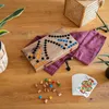 A wooden Tock game board with colorful playing pieces and cards laid out on a wooden table, next to a woven basket and a plant. Produktbild-Vorschau