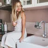 A woman is sitting on the edge of a kitchen sink, holding a glass of water. A water filter is attached to the faucet. Produktbild-Vorschau