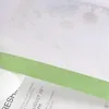 A close-up view of a green ribbon layered over a white fabric with a floral pattern, with text visible on a paper underneath. Produktbild-Vorschau