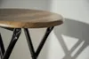 A close-up shot of the top of a stool with a wooden seat and black metal legs, casting a shadow on a white wall. Produktbild-Vorschau