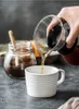 Coffee being poured from a carafe into a mug, with a jar of coffee grounds in the background. Produktbild-Vorschau