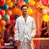 A man in a lab coat stands in front of a backdrop of balloons and streamers, smiling at the camera. Other people are visible in the background. Produktbild-Vorschau
