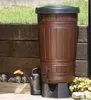 A brown rain barrel with a black lid and a black watering can next to it, placed outdoors in a garden setting with wooden retaining walls and plants. Produktbild-Vorschau