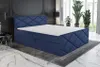A bedroom scene with a blue upholstered boxspring bed as the main focus, placed on a rug with a marble-patterned floor. The background includes decorative wall molding, a side table with a lamp, and large potted plants near a window. Produktbild-Vorschau