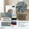 A folded grey fitted sheet is shown in the foreground, with a stack of other colored sheets behind it. A washing machine is visible in the background. Close-up of fabric texture is shown in a circle at the top right. Text describes care instructions. Produktbild-Vorschau