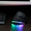 A black vertical gaming mouse with RGB lighting is placed on a wooden desk next to a keyboard, with a monitor in the background. Produktbild-Vorschau