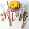 A collection of colorful whisks with wooden handles arranged on a marble surface, with a bowl of whisked eggs in the background. Produktbild-Vorschau