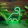 A green neon dinosaur sign is displayed on a wooden wall, surrounded by lush green plants and artificial grass. Podgląd zdjęcia produktu