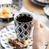 A hand holding a coffee mug with a black and white pattern, with breakfast items in the background. Náhled obrázku produktu