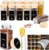A collection of glass spice jars with wooden lids and labels, along with accessories like a funnel and brush, arranged on a white background. Náhled obrázku produktu