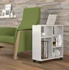 A white rolling side table with a laptop, cup, hourglass, books, and decorative spheres is placed next to a green armchair in a room with a stone-tiled wall and wooden floor. Náhled obrázku produktu