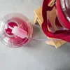 A close-up overhead view of a large glass carboy filled with pink liquid, connected to a siphon tube. A red fabric strap is draped over a wooden stand next to the carboy. Produktbild-Vorschau