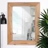 A rectangular mirror with a wooden frame is shown against a white wall. A floor lamp and a plant are visible to the left and right of the mirror, respectively. The mirror reflects a window and a bookshelf. Produktbild-Vorschau