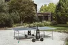 An outdoor ping pong table is set up on a gravel surface in a garden with trees and modern buildings in the background. Produktbild-Vorschau