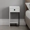 A white bedside table with a black metal leg is shown next to a bed. Podgląd zdjęcia produktu