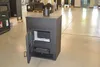 A dark gray wood-burning stove with its door open, displayed in a showroom or warehouse setting. Produktbild-Vorschau