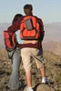 Two people with backpacks are standing on a rocky outcrop, looking out at a mountainous landscape. Produktbild-Vorschau
