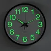 A close-up view of a round, wall-mounted clock with glowing green numbers and hands. Voir les images du produit