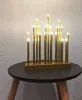 A gold LED candle lamp with 19 flames sits on a dark round table with wooden legs, against a textured light gray and white wall. Produktbild-Vorschau