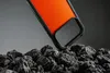 A close-up shot of an orange phone case with a black rim, resting on a pile of dark, rough rocks. Podgląd zdjęcia produktu