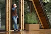 A man stands on a wooden deck outside a modern wooden house, holding a cup and looking out. Produktbild-Vorschau