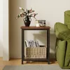 A square side table with a wooden top and metal frame, holding a vase of flowers, a candle, and a small radio. A wicker basket filled with magazines is on the lower shelf. A green sofa is visible to the right. Produktbild-Vorschau