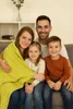 A family of four is sitting on a couch, with the mother holding a green blanket. Produktbild-Vorschau