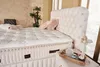 A bedroom scene featuring a bed with a tufted headboard, mattress, and decorative pillows, along with a small table with decorative items. Produktbild-Vorschau