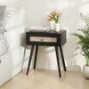 A black nightstand with a rattan drawer front is placed next to a bed. A vase with yellow flowers sits on top of the nightstand. Podgląd zdjęcia produktu