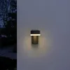 A modern outdoor wall light is mounted on a textured wall at dusk. Produktbild-Vorschau