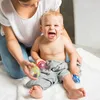 A mother is playing with her baby, who is sitting on a white towel and holding rattles. Produktbild-Vorschau