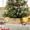 A Christmas tree with decorations and lights, placed on a wooden tree collar, with gifts and a Santa figurine on the floor. Produktbild-Vorschau
