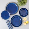 A collection of blue ceramic plates and bowls arranged on a light gray surface, with two lemon halves and a small glass of lemon water in the foreground, and a green plant in the background. Produktbild-Vorschau