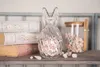 A transparent glass bunny-shaped candy dish filled with colorful marshmallows sits on a shelf next to a stack of decorative books and a glass jar of marshmallows. Produktbild-Vorschau