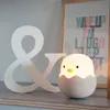 A cute chick-shaped lamp sits on a shelf next to a large ampersand decoration. A framed poster with text is in the background. Produktbild-Vorschau