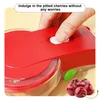 A close-up shot of a red cherry pitter with a hand pressing a button, and a cluster of cherries in the lower right corner. Náhled obrázku produktu