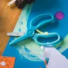 A pair of blue-green training scissors is placed on a blue paper with green and white paint strokes. There is a small purple paint splatter next to the scissors. Some cut white paper is visible in the foreground. Voir les images du produit