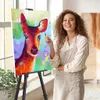 A woman is painting a colorful deer on a canvas placed on a tripod easel in a bright room. Podgląd zdjęcia produktu